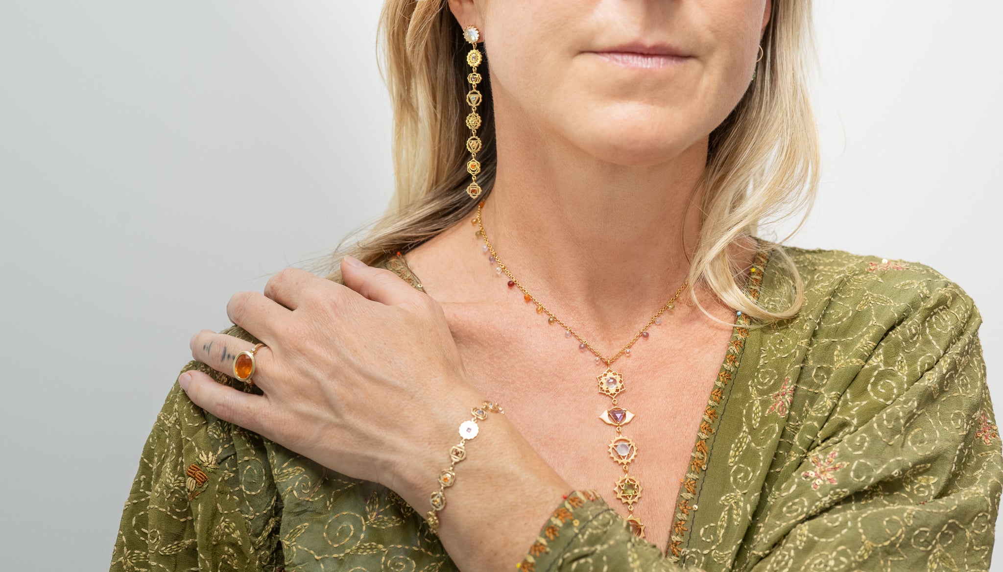 Frau trägt goldenen Karma-Schmuck mit allen sieben Chakren – filigrane Halskette, Armband und Ohrringe in Gold mit Edelsteinen, Granat, Karneol, Citrin, Peridot, Chalcedon, Amethyst, Bergkristall, inspiriert von spiritueller Energie und Balance.